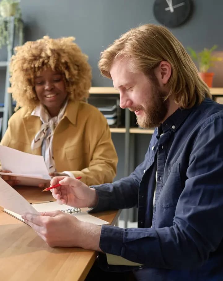 Two people reading printed papers at a meeting table, one making notes in pen.