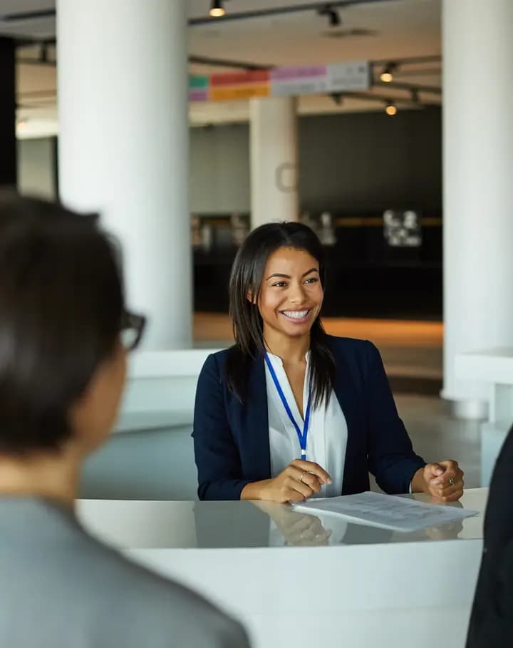 A woman with a lanyard at a registration desk, smiling at two guests as they arrive.